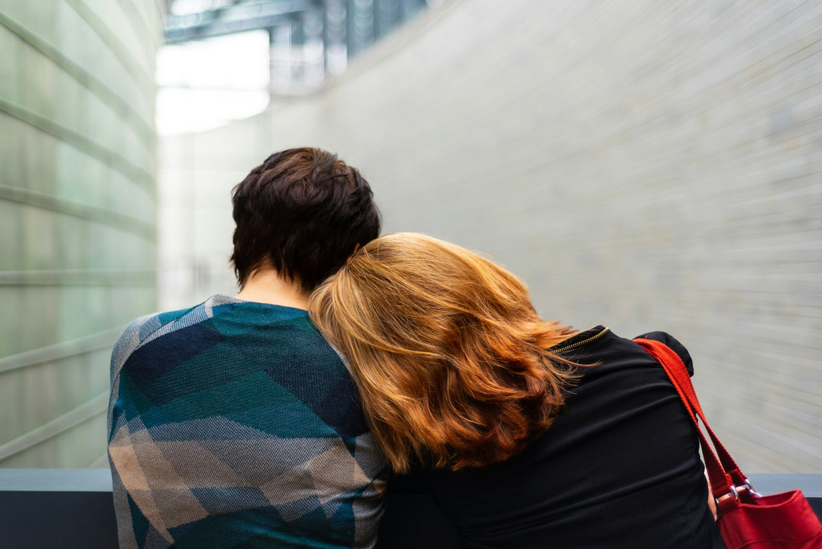 Two people sitting close together, one resting their head on the other's shoulder, seen from behind