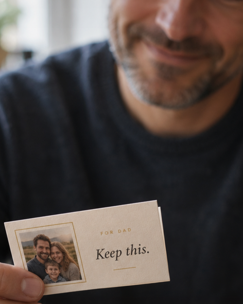 A smiling man holding a small folded Keep This letter reading For Dad, Keep this, with a family photo on the front